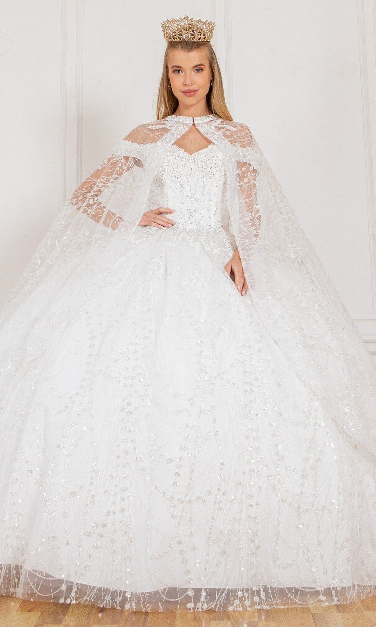 A woman is wearing the Quinceanera Dress 4201 by Cinderella Couture in white, featuring embellishments, a long lace cape, and a gold crown, standing against a plain white background.
